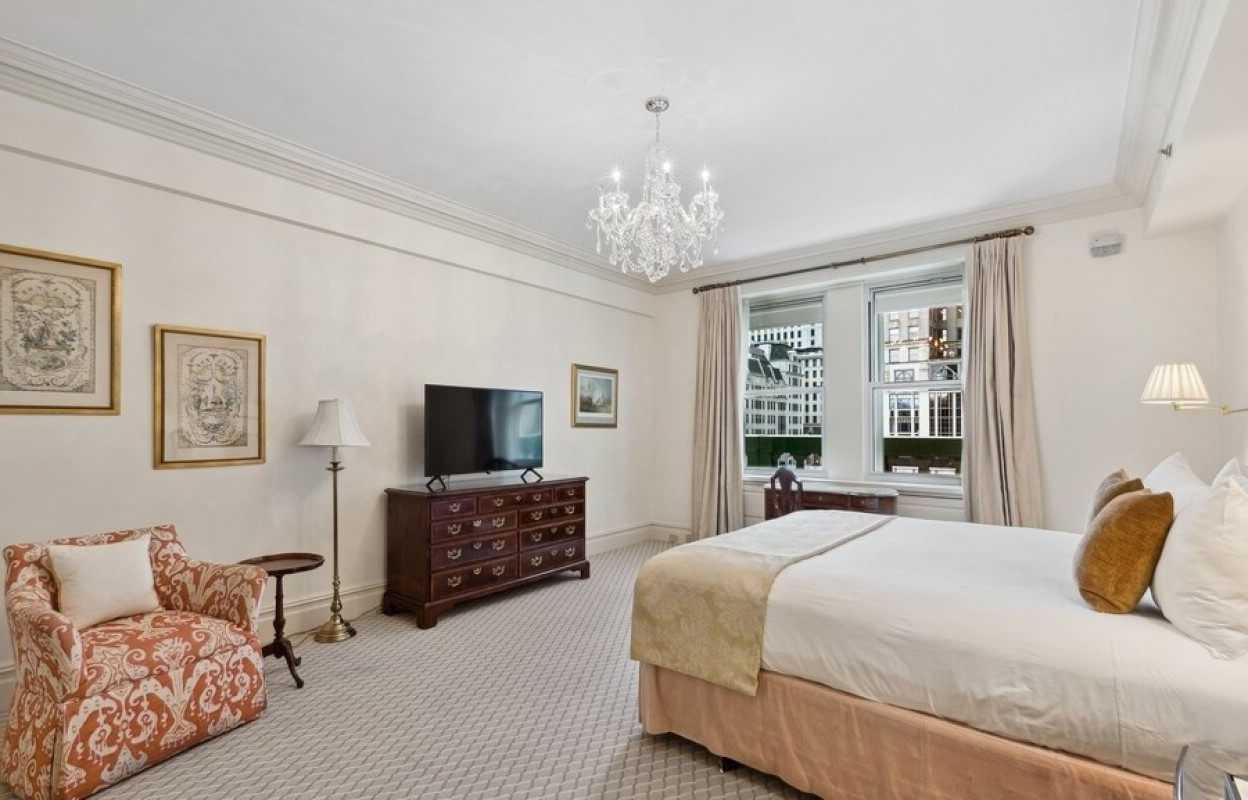 Spacious hotel bedroom with a king-sized bed, crystal chandelier, antique wooden dresser, upholstered armchair, and large windows offering cityscape views.