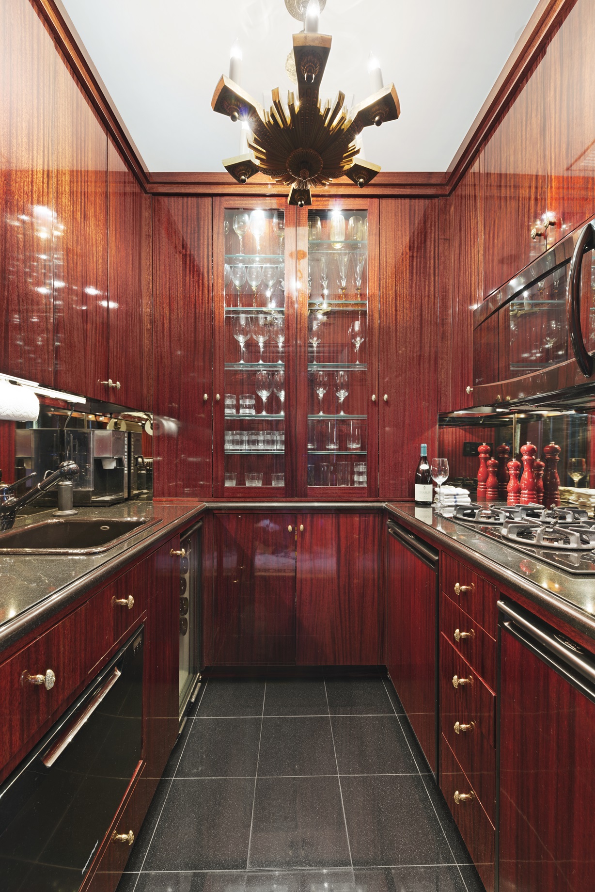 Luxurious galley kitchen with high-gloss mahogany cabinetry, mirrored backsplash, glass-front display shelves filled with stemware, black granite countertops, and a mid-century style starburst ceiling light fixture.
