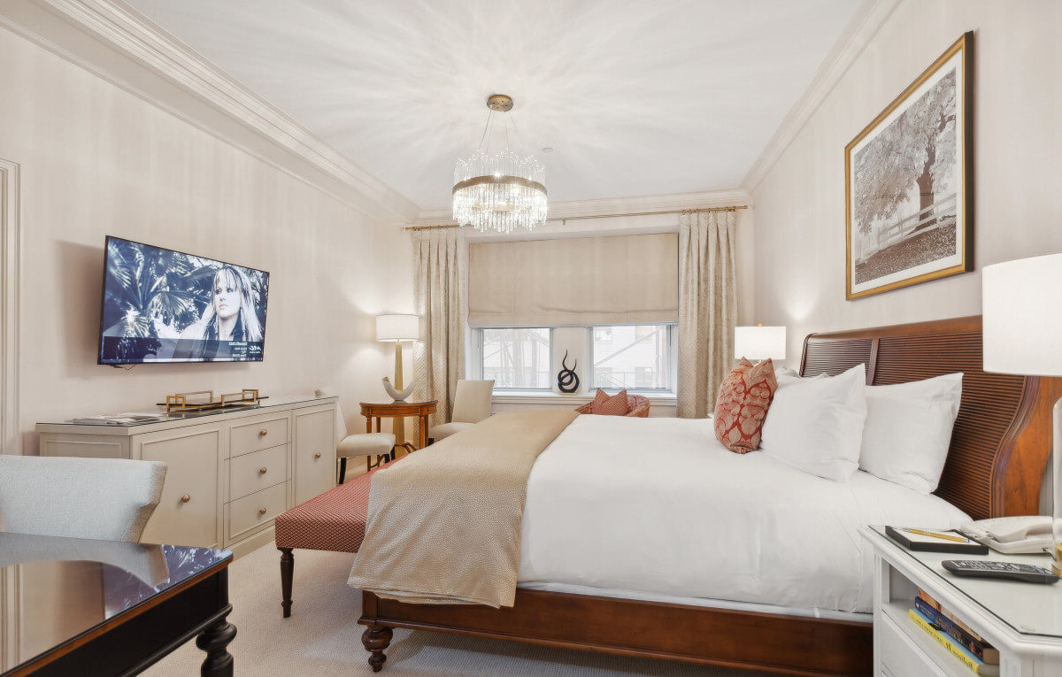 Bright, tastefully furnished hotel room with a king bed, crystal chandelier, flat-screen TV, and traditional decor featuring warm neutrals and rich wood tones.