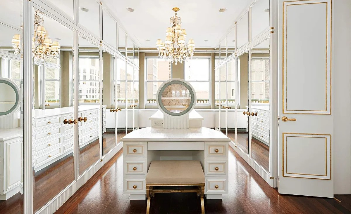 A spacious and luxurious walk-in closet at The Sherry-Netherland, featuring mirrored doors, ample storage, a vanity table with a stool, and an elegant chandelier.