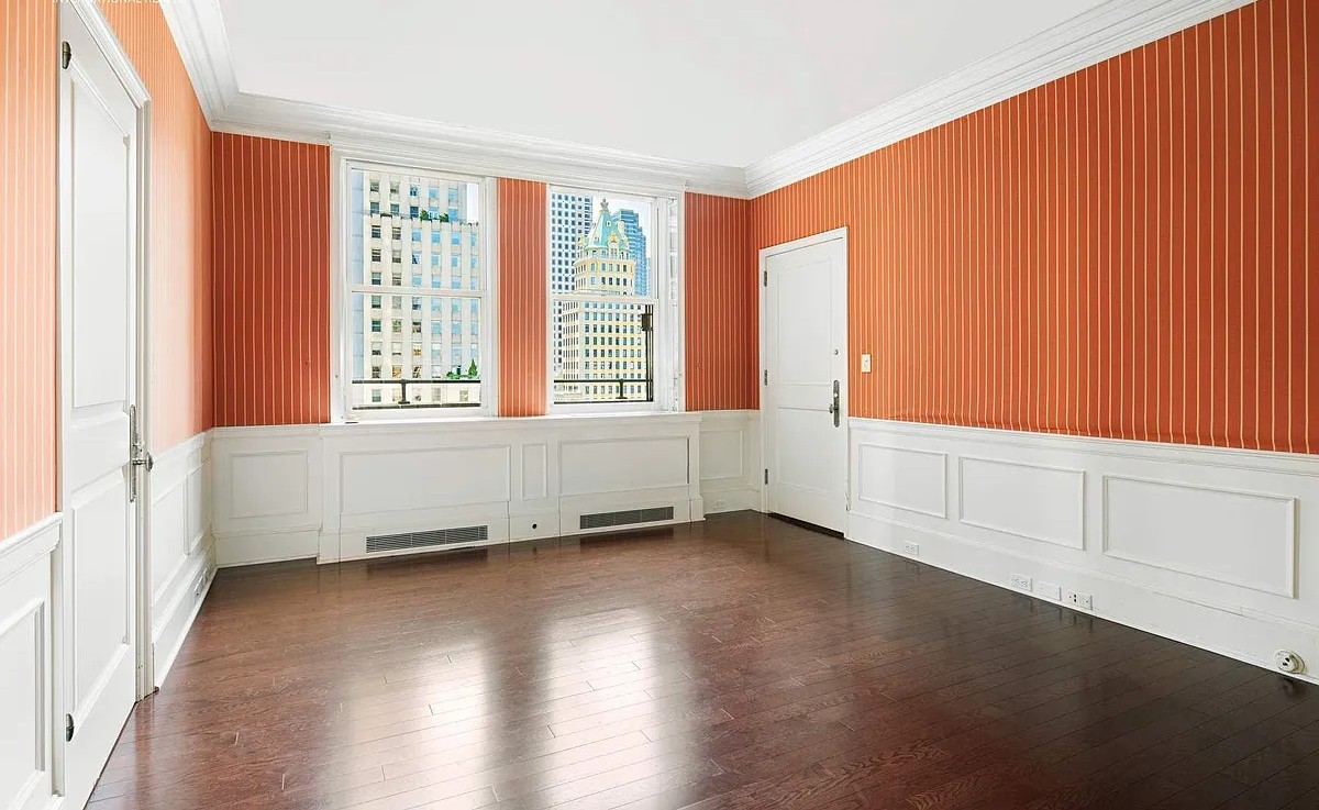 An empty room at The Sherry-Netherland featuring vibrant orange and white striped wallpaper, dark hardwood floors, and two large windows offering city views.