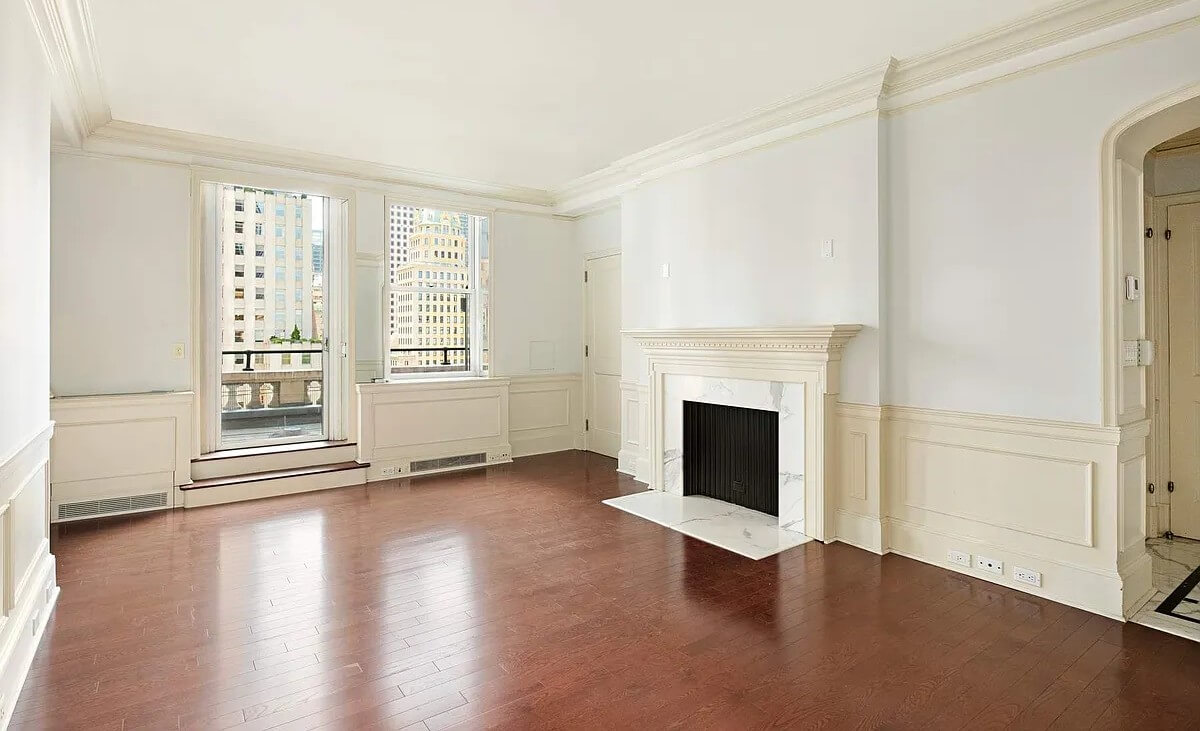 An empty, light-filled room at The Sherry-Netherland featuring a white marble fireplace, dark hardwood floors, wainscoting, and French doors leading to a balcony with city views.