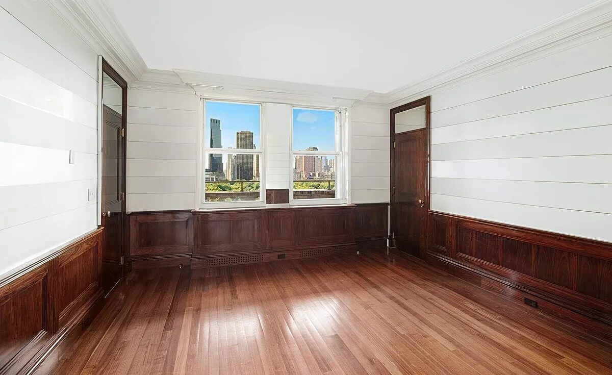 An unfurnished room at The Sherry-Netherland featuring rich dark wood paneling on the lower walls, white horizontal paneling above, hardwood floors, and two windows with city views.