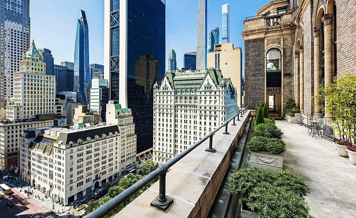An elegant terrace with lush greenery and wrought-iron railings, overlooking the iconic Plaza Hotel and the surrounding New York City skyline on a sunny day.