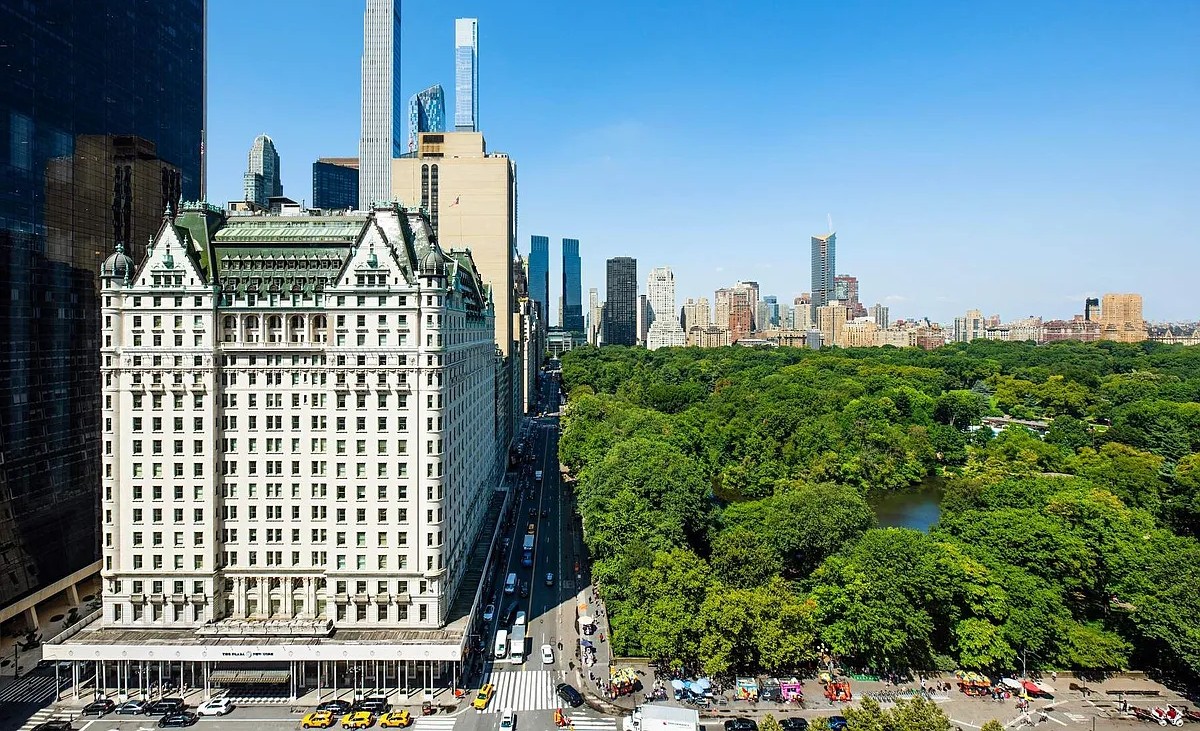 An elevated view of The Plaza Hotel, situated at the southwest corner of Central Park South, with Central Park's greenery and the Midtown Manhattan skyline extending into the distance under a clear blue sky.
