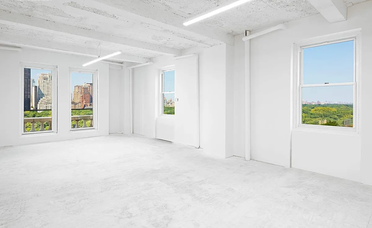 A spacious, all-white empty room with large windows revealing views of the Manhattan skyline and Central Park's greenery.