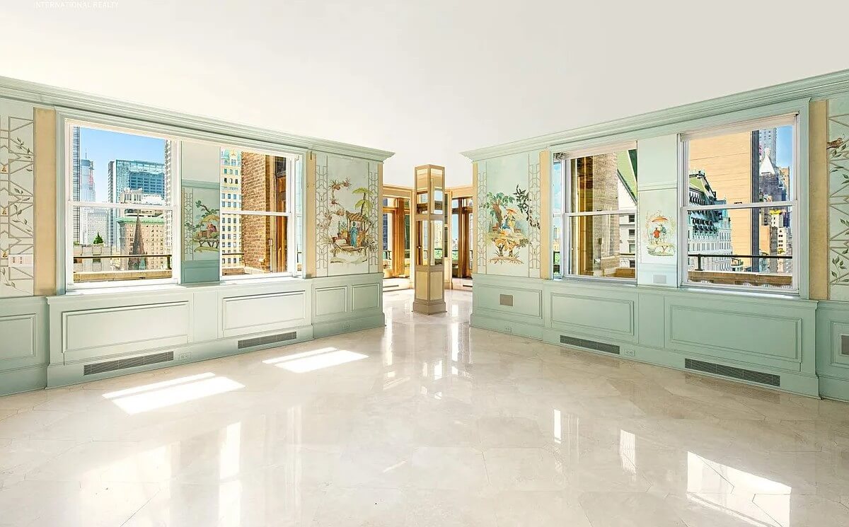 A spacious, empty room at The Sherry-Netherland with light green walls featuring hand-painted decorative panels and large windows offering urban views.
