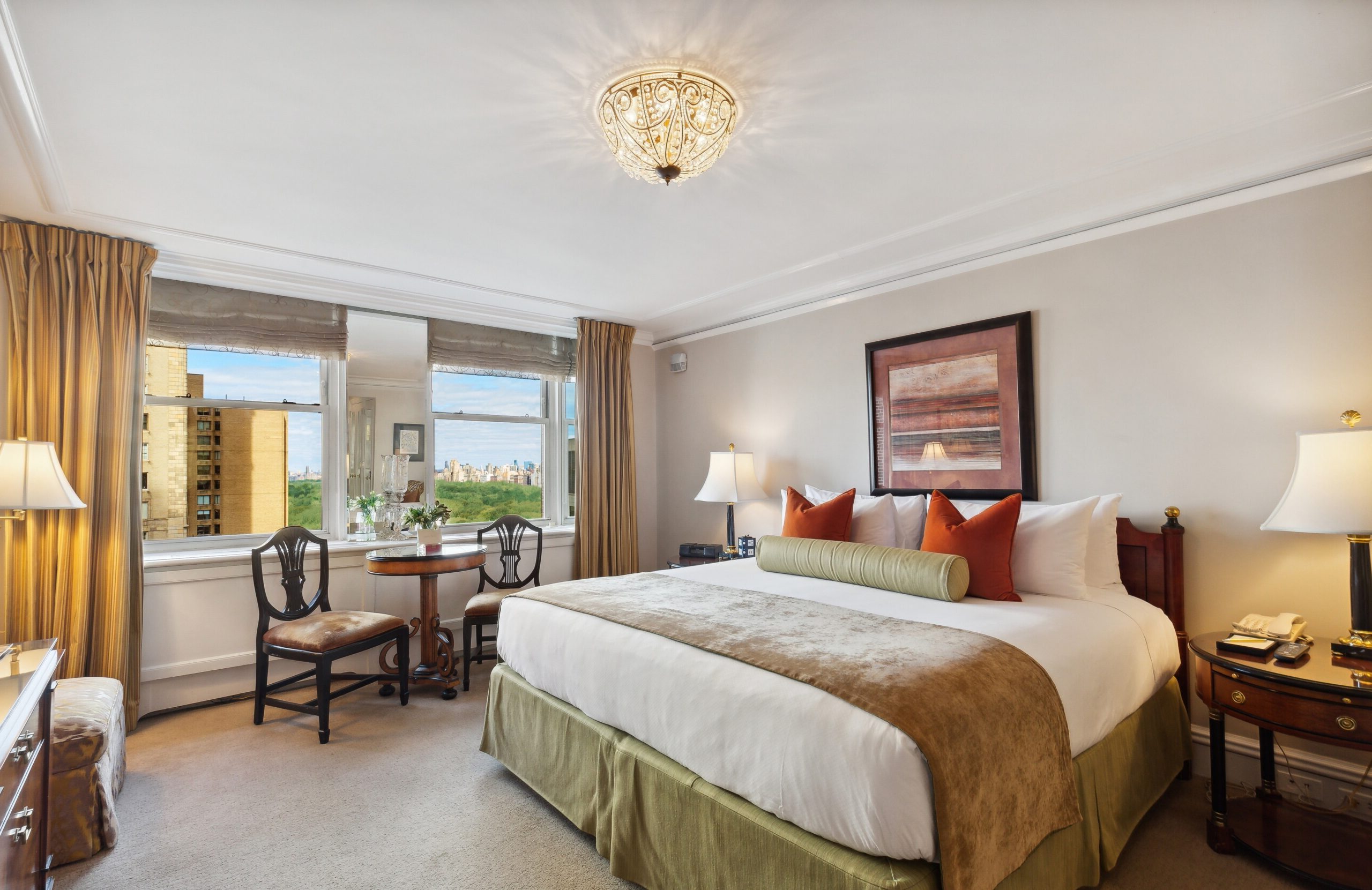 Sunlit hotel room with a king bed, bold accent pillows, classic wood furnishings, and panoramic windows overlooking Central Park and the city skyline.