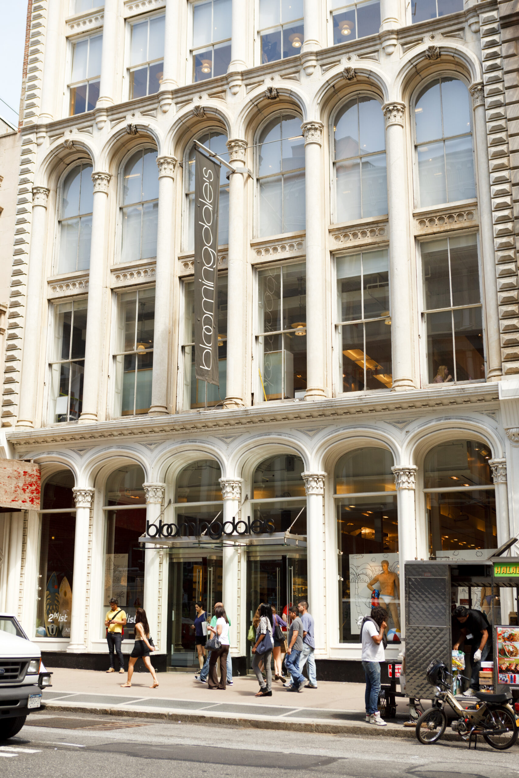 Shoppers walking past the entrance of Bloomingdale’s SoHo in New York City, with a food cart and historic building facade