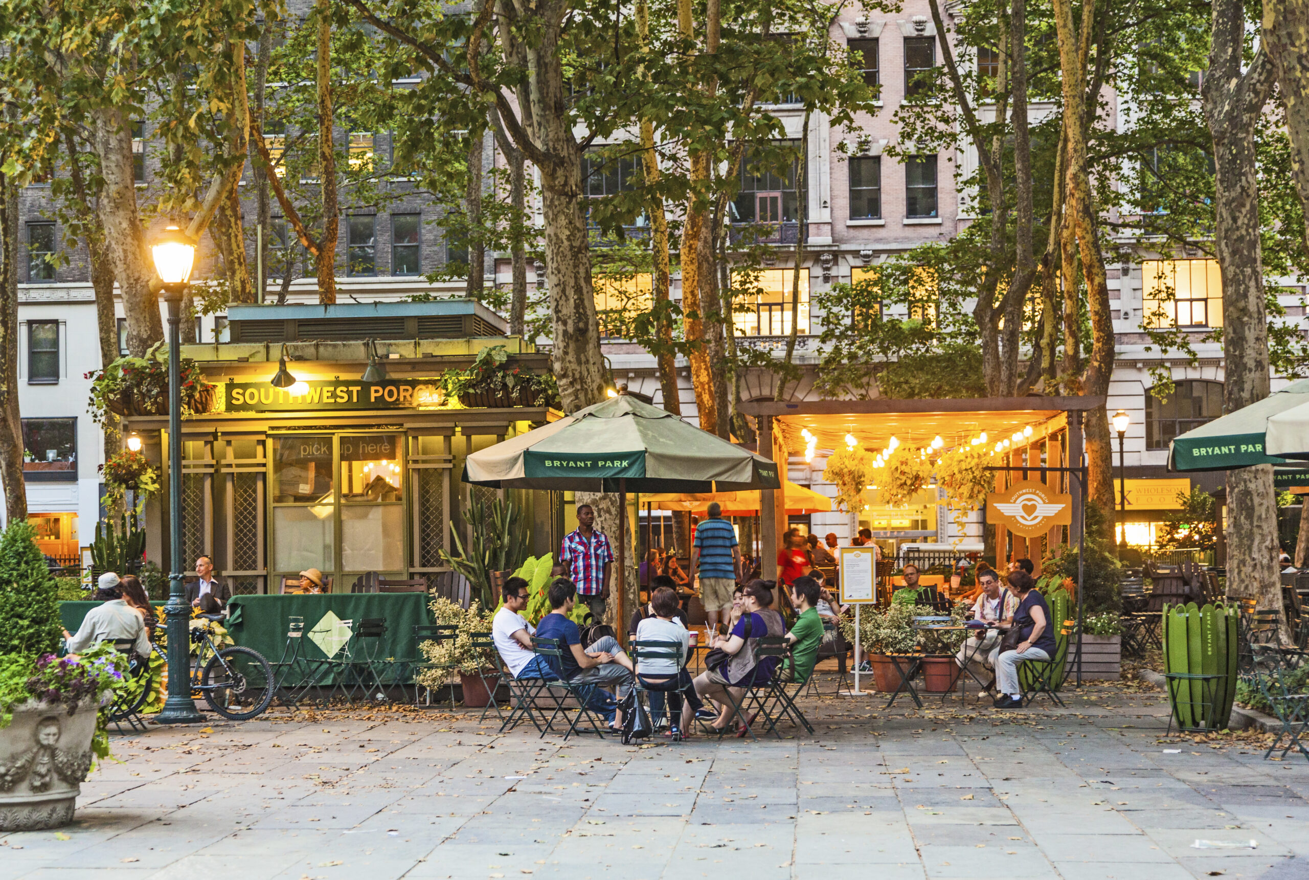 People relaxing at outdoor tables under string lights at Bryant Park in New York City during a warm evening.