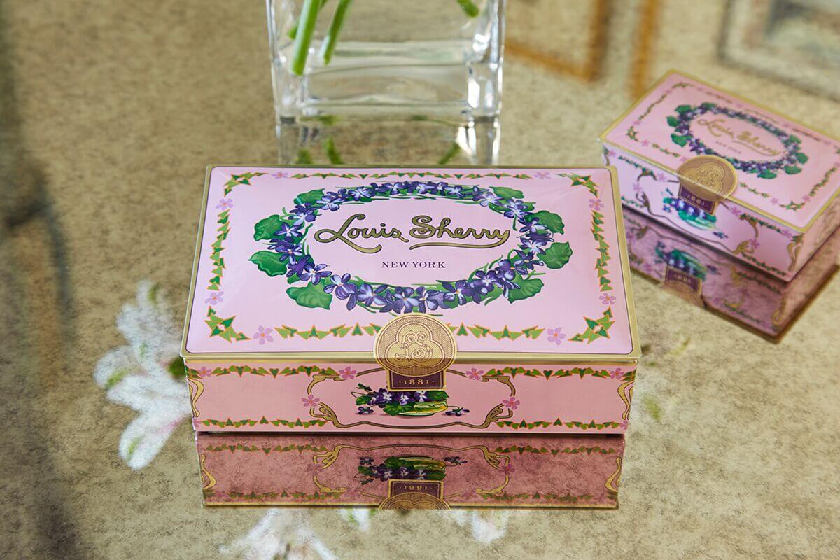 Close-up of a pink Louis Sherry chocolate tin featuring a floral wreath design and gold accents, placed on a marble surface.