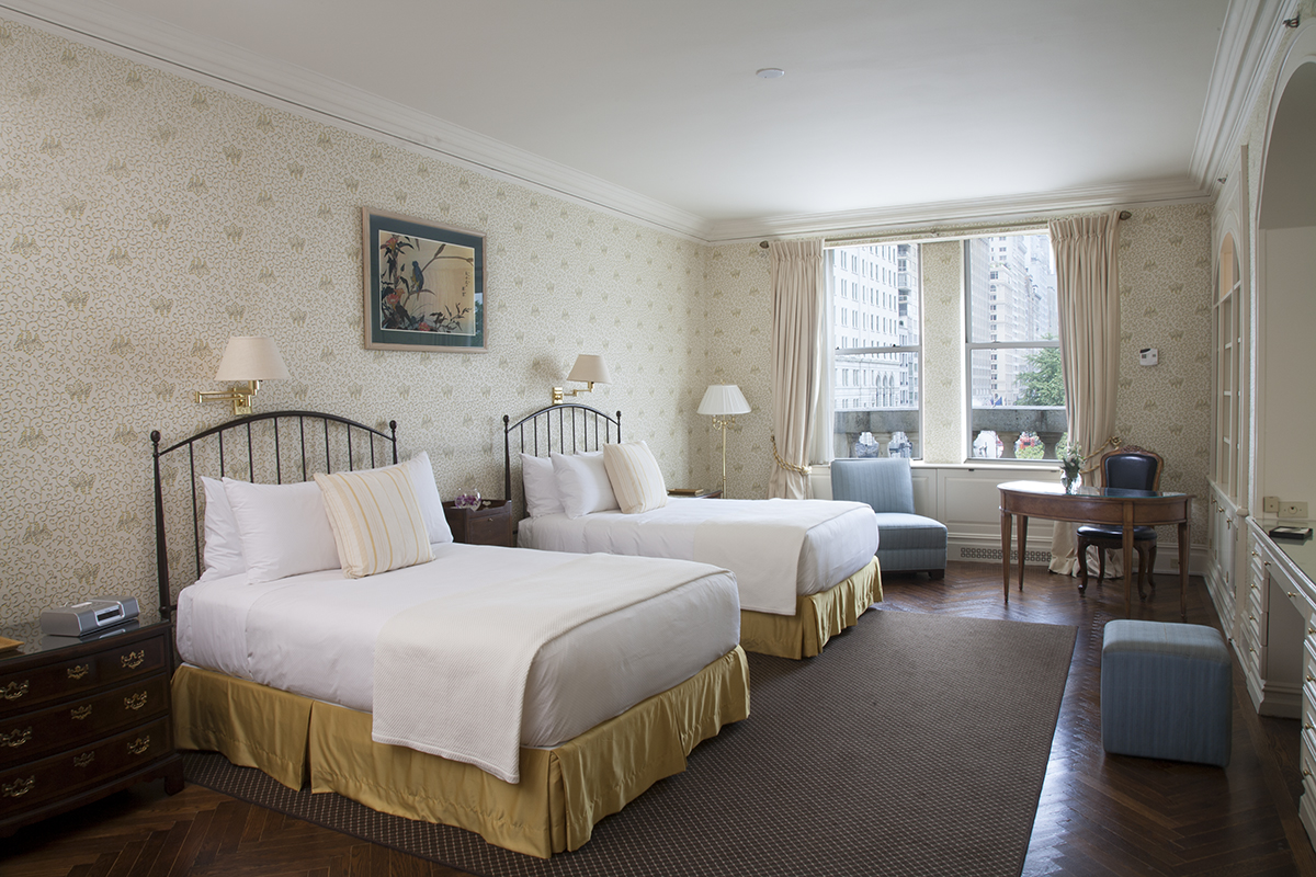 Twin bedroom with herringbone wood floors, floral wallpaper, white linens with gold accents, and city views at the Sherry-Netherland hotel in New York City.